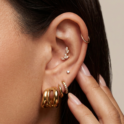 Close-up of a person's ear adorned with multiple gold ear piercings, including hoop earrings, a floral stud, a small dot stud, and stacked huggie hoop earrings, with a hand gently touching the ear.