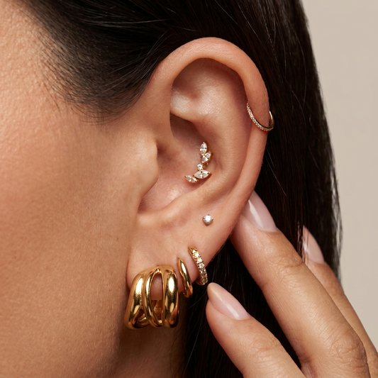 Close-up of a person's ear adorned with multiple gold ear piercings, including hoop earrings, a floral stud, a small dot stud, and stacked huggie hoop earrings, with a hand gently touching the ear.